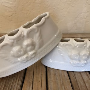 May include: Two white ceramic toothbrush holders with a decorative design of cherubs. Each holder has three slots for toothbrushes.