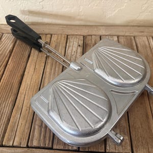 May include: A vintage silver-colored sandwich maker with a black handle. The clamshell-style cooker features a design with radiating lines. The handle is made of black plastic and is attached to the metal cooking surface with metal rods.