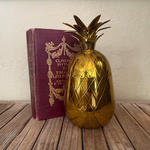 May include: A gold pineapple-shaped ice bucket with a textured surface. The bucket is sitting on a wooden surface next to a book with a burgundy cover.