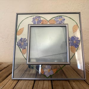 May include: A square glass picture frame with a silver metal border. The frame features a heart-shaped design with blue flowers and tan leaves. A smaller square frame is centered within the larger frame.