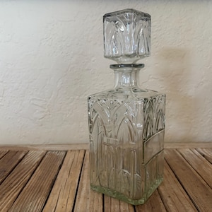 May include: Clear glass decanter with a square base and stopper. The decanter features a decorative, etched design on the front and sides. The stopper is also square and has a matching etched pattern. The decanter is sitting on a wooden surface.
