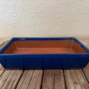 May include: A rectangular, blue ceramic planter with a terracotta interior. The planter has a glossy finish and is set on a wooden surface. The design is simple and modern, suitable for small plants or succulents.