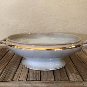 May include: A white ceramic serving bowl with gold trim and two handles. The bowl is on a wooden surface.