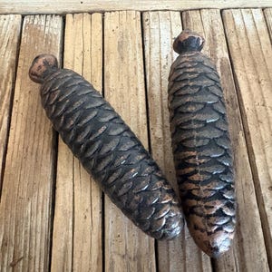 May include: Two elongated, dark brown, metallic pine cone-shaped weights with a textured, scale-like surface. Each object features a small, rounded knob at the top and a tapered, rounded base, displaying a weathered, rustic finish throughout.