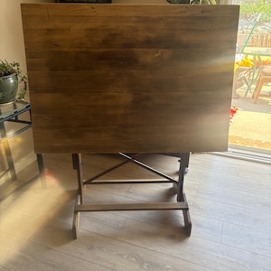May include: A rectangular wooden drafting table with a smooth, medium-brown surface featuring visible wood grain. The table is supported by a sturdy, dark-toned wooden frame with an X-shaped cross-brace structure underneath the main tabletop.