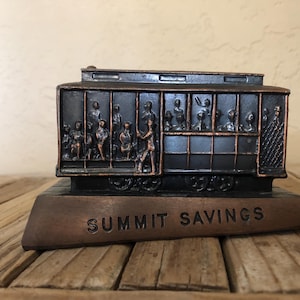 May include: A vintage metal coin bank shaped like a streetcar with a black and copper finish. The streetcar is filled with small figures and the base reads "Summit Savings".