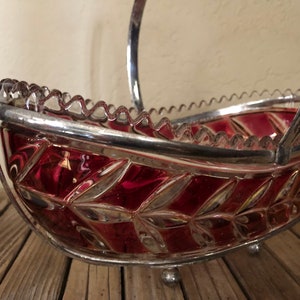 May include: A red glass bowl with a silver rim and handle. The bowl has a decorative pattern of leaves and is sitting on a wooden surface.