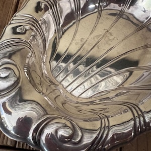 May include: A silver-colored, decorative dish with a shell-like design. The dish has a scalloped edge and a series of curved lines radiating from the center. The surface reflects light, creating highlights and shadows.
