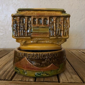 May include: A ceramic music box shaped like a San Francisco cable car with the words "Powell & Hyde" and "San Francisco" on the side. The base of the music box features a depiction of the Golden Gate Bridge.