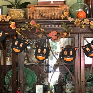 May include: A black and orange Halloween garland with black cat faces hanging from a dark wood cabinet. The garland is decorated with fall leaves.