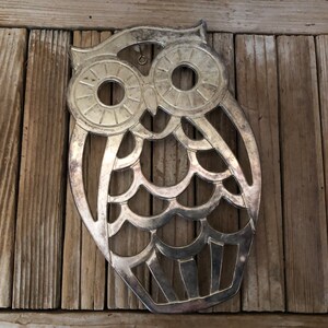 May include: Silver metal owl-shaped hot pad with a cut-out design. The owl has large round eyes and a detailed feather pattern.