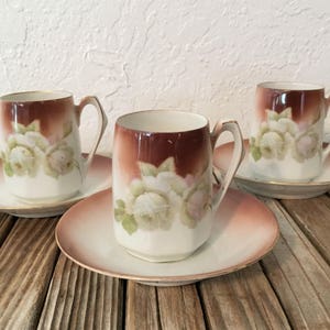 May include: Three white porcelain teacups with brown floral designs and gold trim. Each cup is on a matching saucer with a brown rim.