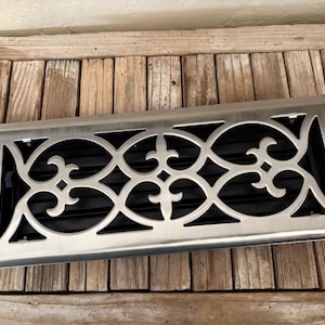 May include: A rectangular metal vent cover with a decorative, openwork design. The cover has a brushed metal finish and a black interior. The design features a repeating pattern of fleur-de-lis and circular shapes.