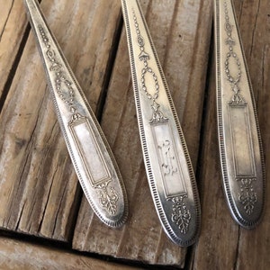 May include: Three silver spoons with ornate designs and a blank rectangular space for engraving. The spoons are laid out on a wooden surface.