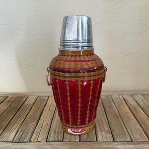 May include: A vintage cocktail shaker with a woven red and tan basket-like body and a silver metal top. The shaker has a cylindrical shape with a wider base and a narrower top. It features small handles and a decorative design.