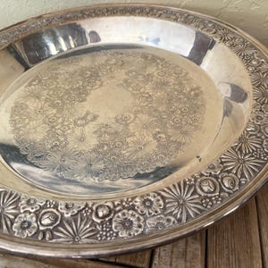 May include: A silver-colored, round serving tray with a floral design. The center of the tray features a delicate pattern of small flowers, while the rim is adorned with a repeating pattern of stylized flowers and leaves. The tray is likely made of metal.