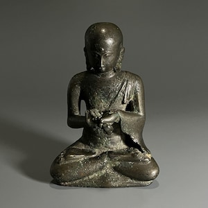 Puede incluir: Estatua de bronce de una figura sentada en posición meditativa. La figura tiene la cabeza rapada, túnicas drapeadas y las manos juntas. La estatua tiene un aspecto envejecido y desgastado con pátina verde.