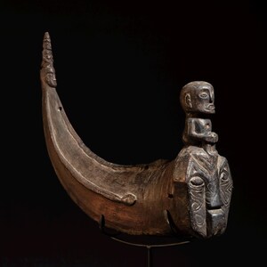 May include: A dark brown, carved wooden sculpture resembling a boat. It has a stylized face at the front and a small figure seated on top. The curved shape and detailed carvings suggest a cultural artifact, possibly of historical significance.