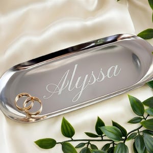 May include: An oval-shaped silver-toned metal tray with a polished reflective surface. The name Alyssa is engraved in a cursive script font in the center of the tray. Two plain gold-toned metal rings are resting on the left side of the tray.