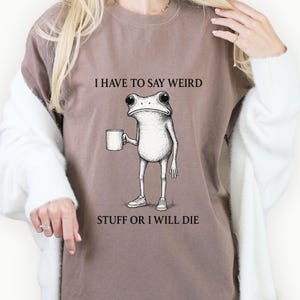 Puede incluir: Camiseta marrón con el texto "I HAVE TO SAY WEIRD STUFF OR I WILL DIE" y una ilustración en blanco y negro de una rana sosteniendo una taza. La rana lleva zapatillas.