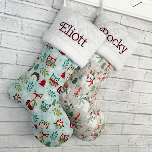 May include: Two Christmas stockings with white quilted cuffs featuring the embroidered names Eliott and Rocky. The left stocking has a light blue background with owls, foxes, and snowflakes. The right stocking has a beige background with polar bears, gnomes, and trees.