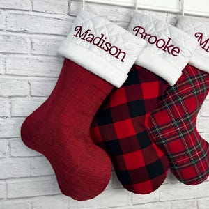 May include: Three Christmas stockings with white quilted cuffs featuring the embroidered names Madison, Brooke, and Mia in red thread. The first stocking is solid red, the second has a red and black buffalo check, and the third has a red plaid.