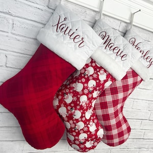 Peut inclure: Trois chaussettes de Noël suspendues en rangée. La première est à carreaux rouges, la deuxième présente un fond rouge avec des bonhommes de neige et des flocons blancs, et la troisième est à carreaux rouges et blancs avec des noms brodés.