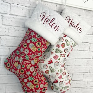 May include: Two Christmas stockings with white quilted cuffs featuring the embroidered names Helen and Nadia in dark red script. One stocking has a red background with a cookie pattern, while the other has a white background with Santa Claus motifs.