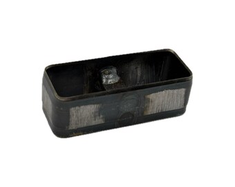 May include: A rectangular metal cutting die with a hollow center and rounded corners. The exterior surface features a dark, oxidized finish with visible brushed metallic patches and a central welded joint on the inner wall.