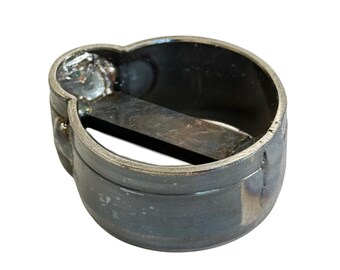 May include: A circular metal ring with a dark, oxidized finish featuring a flat horizontal bar across the center. The exterior surface shows visible weld marks and a slightly uneven texture, with a small protruding section on one side of the band.