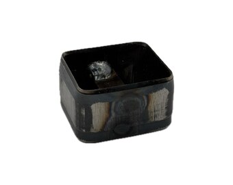 May include: A square-shaped metal die cutter with a dark, oxidized finish and visible weld marks on the exterior sides. The interior features a small, metallic skull-shaped emblem attached to a flat metal bar spanning the center of the frame.