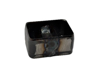 May include: A rectangular metal component with rounded corners, featuring a dark, heat-treated finish. The exterior surface displays visible grinding marks and metallic textures, with a hollow center and a small welded metal tab inside.