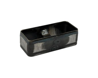 May include: A rectangular metal component with a dark, unfinished surface featuring visible grinding marks and a central welded bridge. The object has rounded corners and an open top, showing a raw industrial texture with metallic gray tones.