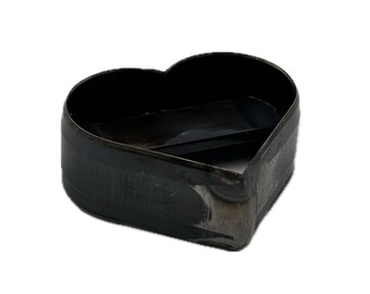 May include: A heart-shaped metal cutting die with a dark, oxidized finish. The tool features a continuous, sharp-edged wall forming a symmetrical heart outline, with a hollow center and a flat, open base, showing signs of industrial wear.