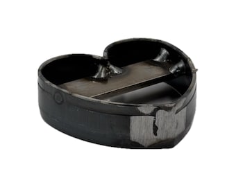 May include: A heart-shaped metal cutter with a dark, oxidized finish. The tool features a horizontal metal bar welded across the center of the heart frame, with visible weld marks and a small area of silver-toned metal showing through the dark surface.