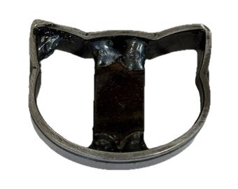 May include: A dark metallic cat-shaped ring featuring a stylized feline head outline with pointed ears. The band is constructed from a continuous metal strip with visible welded joints connecting the ears to the central vertical bar component.