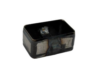May include: A rectangular metal cutting die with a hollow center and a dark, oxidized finish. The exterior walls feature brushed silver-toned metallic patches where the dark coating has been removed, revealing the raw steel underneath.
