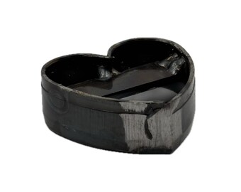 May include: A heart-shaped metal cookie cutter with a dark, oxidized finish. The object features a continuous band of metal formed into a heart outline with a horizontal support bar welded across the center, showing visible weld marks and texture.