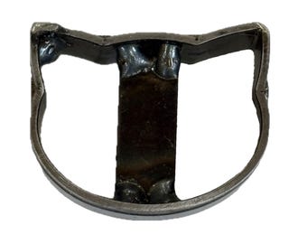 May include: A dark gray metal object shaped like a stylized cat head outline with a vertical bar centered in the middle. The piece features visible welded joints at the connection points between the outer frame and the central vertical metal strip.