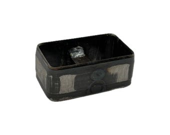 May include: A rectangular metal box with a dark, weathered finish. The exterior features visible brushed metal patches and a central circular indentation. Inside, a small metal tab is welded to the bottom surface of the container.