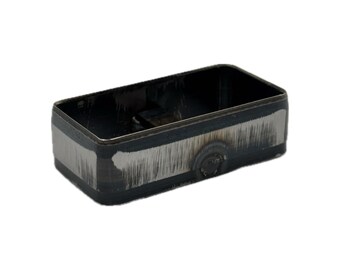 May include: A rectangular metal container with rounded corners, featuring a dark, oxidized finish and visible brushed silver-toned vertical scratch marks along the exterior sides. The interior is hollow and dark, showing a metallic construction.