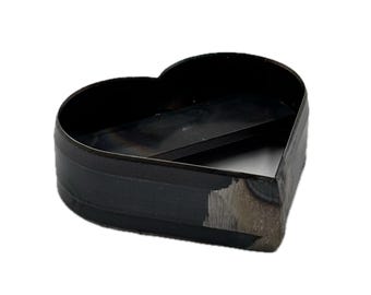 May include: A heart-shaped metal cutting die with a dark, oxidized finish and a visible weld seam on the side. The tool features a sharp, raised edge designed for precision cutting, showcasing a raw industrial appearance with metallic gray tones.