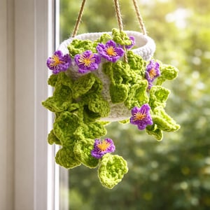 Pode incluir: Um vaso suspenso de crochê feito à mão com um vaso branco, folhas verdes e flores roxas com centros amarelos. O vaso é suspenso por uma corda de fibra natural, criando uma peça decorativa e charmosa.