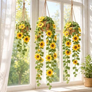 Puede incluir: Tres cestas colgantes de girasoles de ganchillo. Cada cesta está llena de hojas verdes y girasoles amarillos. Los girasoles caen en cascada, creando una exhibición vibrante. Las cestas están suspendidas por cuerdas marrones.