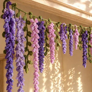 May include: Crocheted wisteria garland in shades of purple and pink, with green leaves. The handmade floral decor hangs from a wooden frame, adding a touch of nature-inspired charm to any space. Perfect for home decoration.