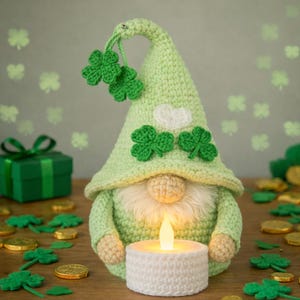 Op de afbeelding: Een lichtgroene gehaakte kabouter met een witte baard en een oplichtende kaars. De hoed van de kabouter is versierd met groene klavers en een witte wolk. Een groene geschenkdoos en gouden munten staan op de achtergrond.