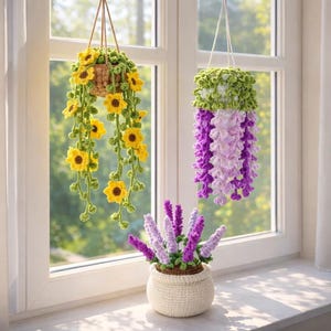 Könnte beinhalten: Gehäkelte Hängekörbe und eine Topfpflanze. Ein Korb zeigt gelbe Sonnenblumen mit grünen Ranken, der andere lila und lavendelfarbene Blumen. Ein kleiner weißer Topf enthält lavendelfarbene Blumen, alles in der Nähe eines Fensters.