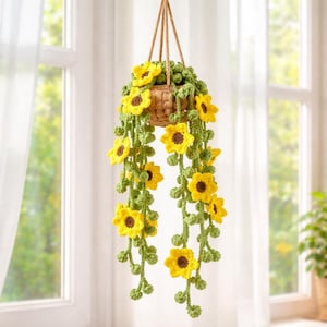 Puede incluir: Una cesta colgante de ganchillo con flores de girasol amarillas y vides verdes. La cesta está hecha de material tejido y suspendida por una cuerda marrón. Los girasoles tienen centros marrones y están rodeados de pétalos amarillos.
