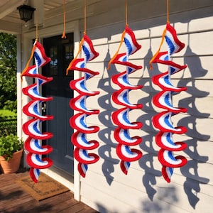 Puede incluir: Cuatro decoraciones espirales de ganchillo colgantes. Cada espiral presenta rayas rojas, blancas y azules, con un diseño similar a una bandera en la parte superior. Las decoraciones están suspendidas de cuerdas naranjas, creando una exhibición festiva y llamativa.