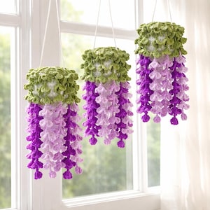 May include: Three hanging crochet flower pot covers in a window. The tops are green, the middle is light purple, and the bottom is dark purple. The crochet design resembles wisteria flowers. The pots are suspended by white cords.